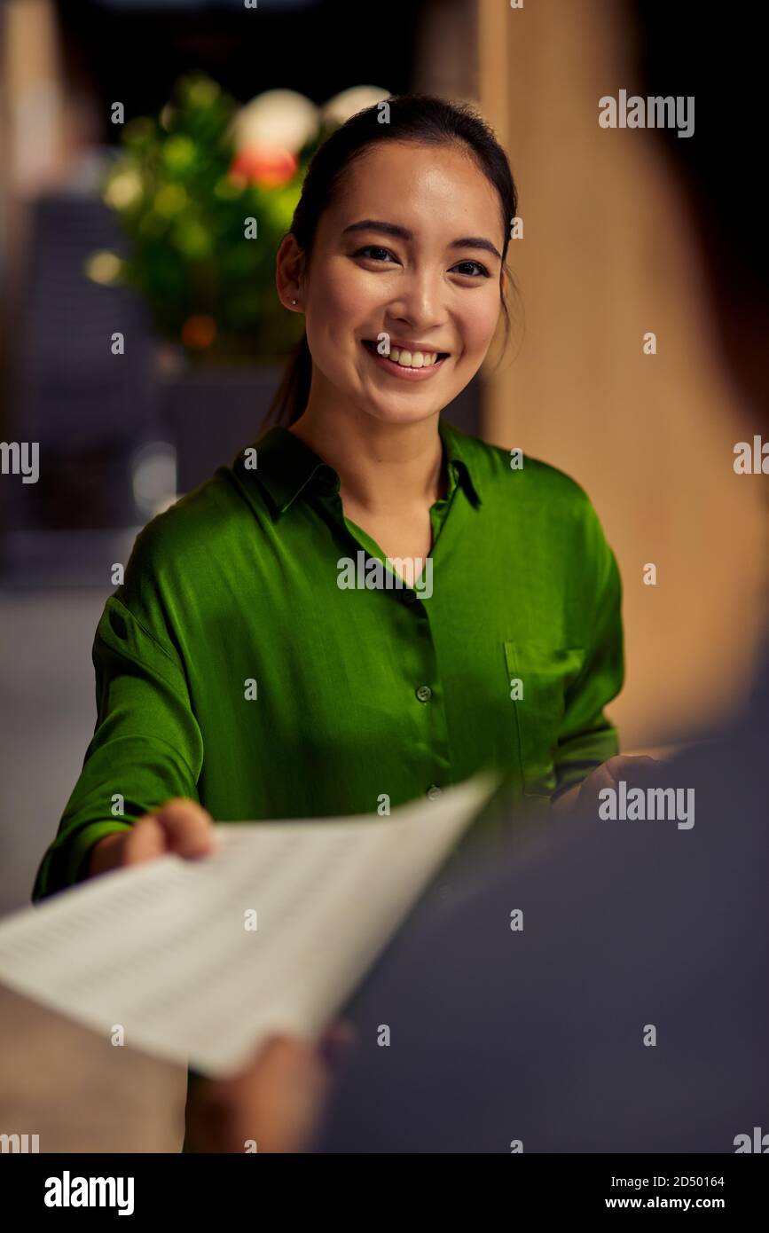 Immagine verticale di una giovane e felice lavoratrice asiatica d'ufficio che dà alcuni documenti alla sua collega e sorridendo mentre si sta insieme nell'ufficio moderno. Business, lavoro di squadra, concetto di cooperazione Foto Stock