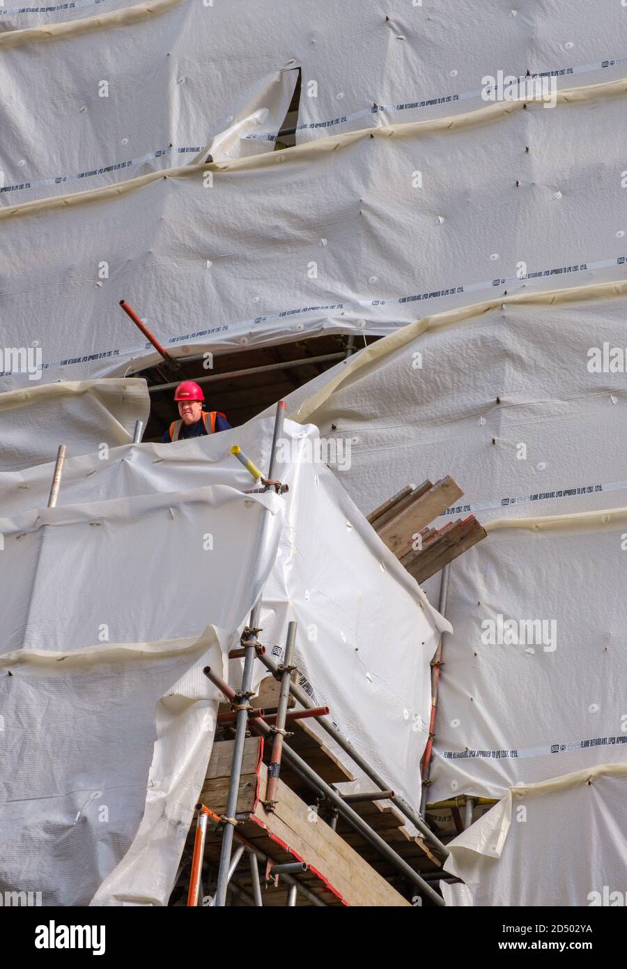 Operaio di costruzione in arancione casco cime fuori foro in copertura del sito e guarda verso il basso. Angolo tra Oxford St e Duke St, Londra, W1, Inghilterra. Foto Stock