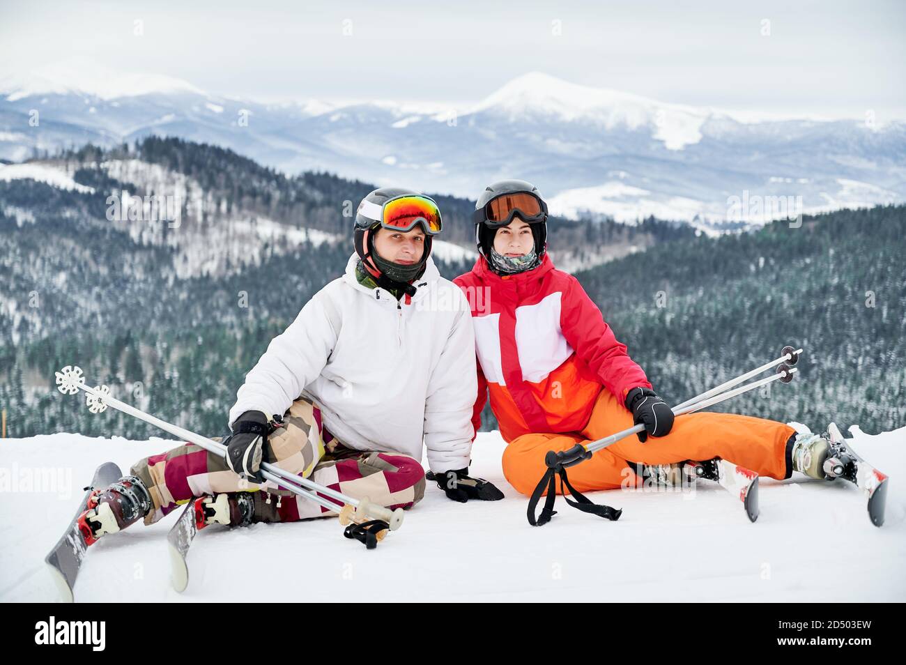 Due amici che indossano abiti da sci e attrezzature da sci si trovano in montagna d'inverno godendo di tempo innevato, sci e paesaggi incredibili. Ritratto di una coppia seduta su una collina innevata che sorride guardando la macchina fotografica Foto Stock