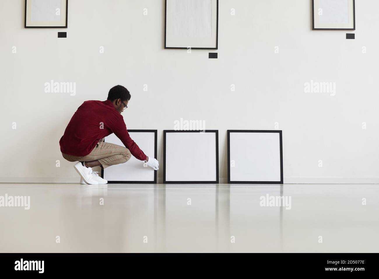 Ritratto grandangolare dell'uomo afro-americano che allestiscono cornici nere vuote sul pavimento mentre pianifica galleria d'arte o mostra, spazio di copia Foto Stock