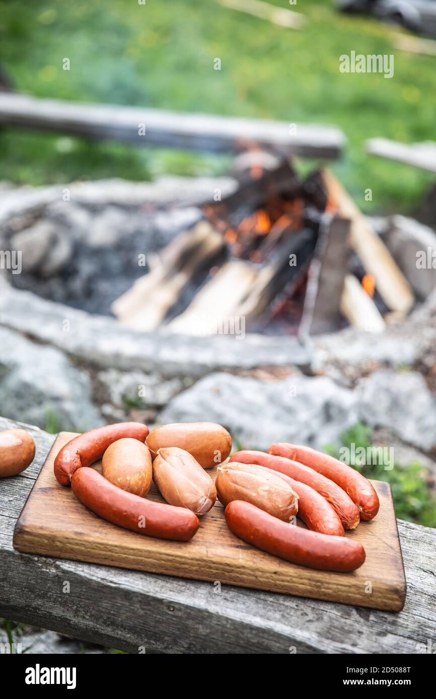 Salsicce su un tagliere di legno pronto per la tostatura un falò sullo sfondo Foto Stock