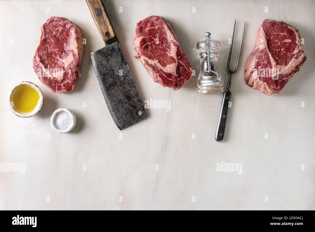 Tre materie non cotte le bistecche di manzo con butcher cleaver, carne forcella, pepe, sale e olio di oliva in fila su marmo bianco dello sfondo. Laici piana, spazio Foto Stock