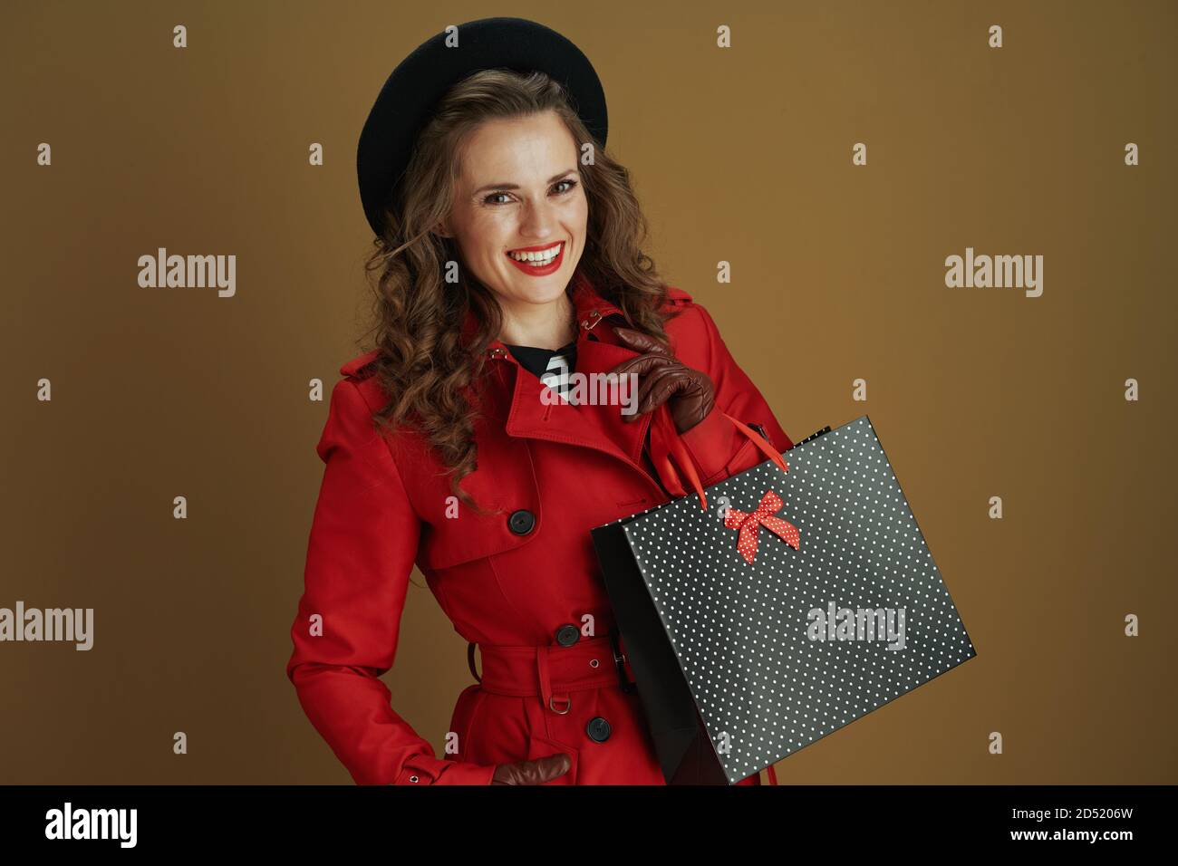 Ciao settembre. Donna trendy felice in cappotto rosso e beretto nero con guanti in pelle e borsa di carta per lo shopping su sfondo marrone. Foto Stock