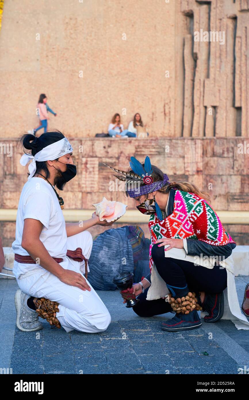 Due partecipanti inginocchiati, uno con copricapo e maschera piumati, offrono una conchiglia in una cerimonia alla Plaza de Colon, Madrid, il 12 ottobre 2020 Foto Stock