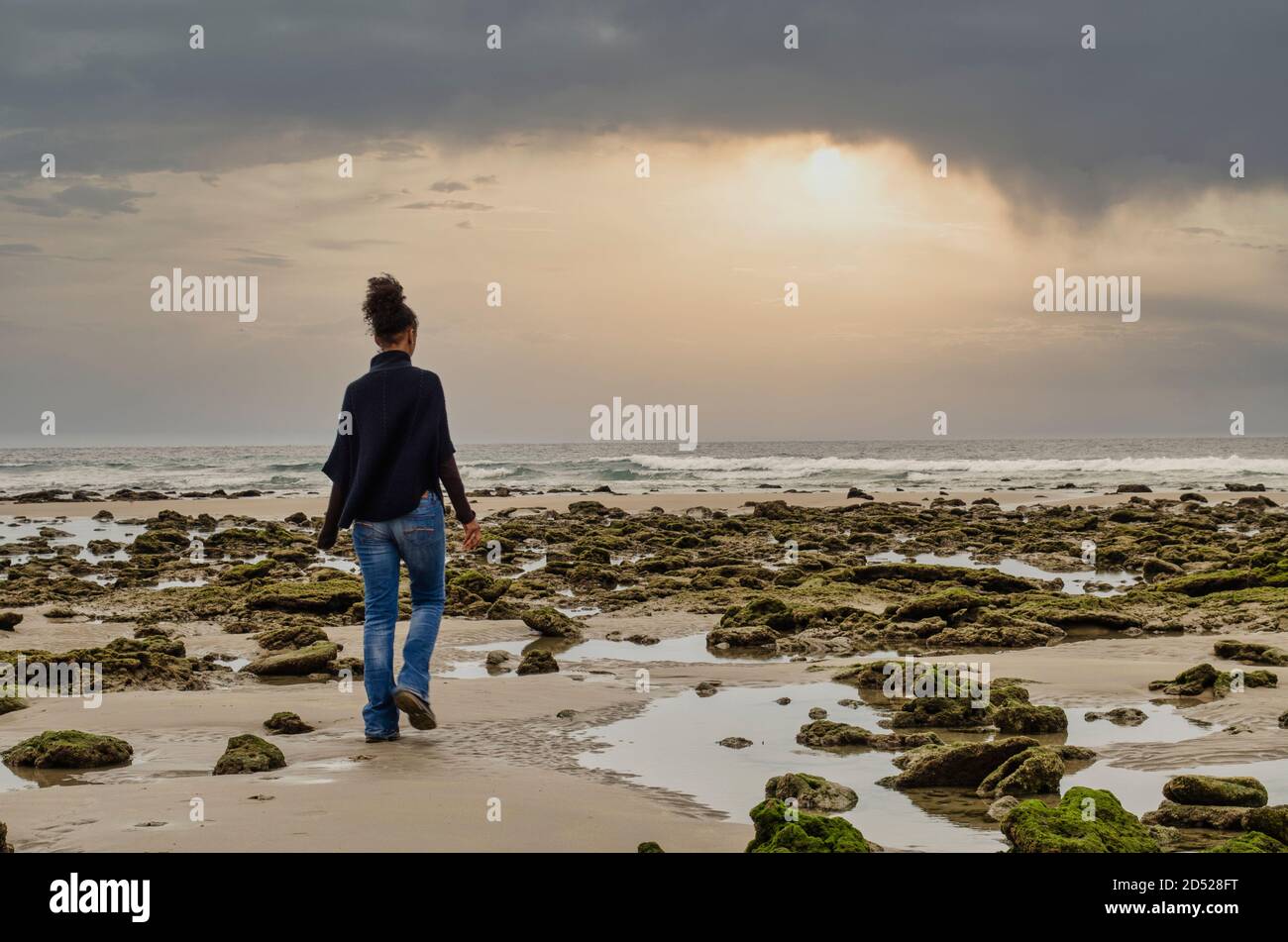 Vista posteriore di una donna nera che cammina sulla spiaggia di Zahora a Cadice tra rocce ricoperte di muschio. Fuoco selettivo sulle rocce. Lungo esposizione.bellissimo tramonto Foto Stock