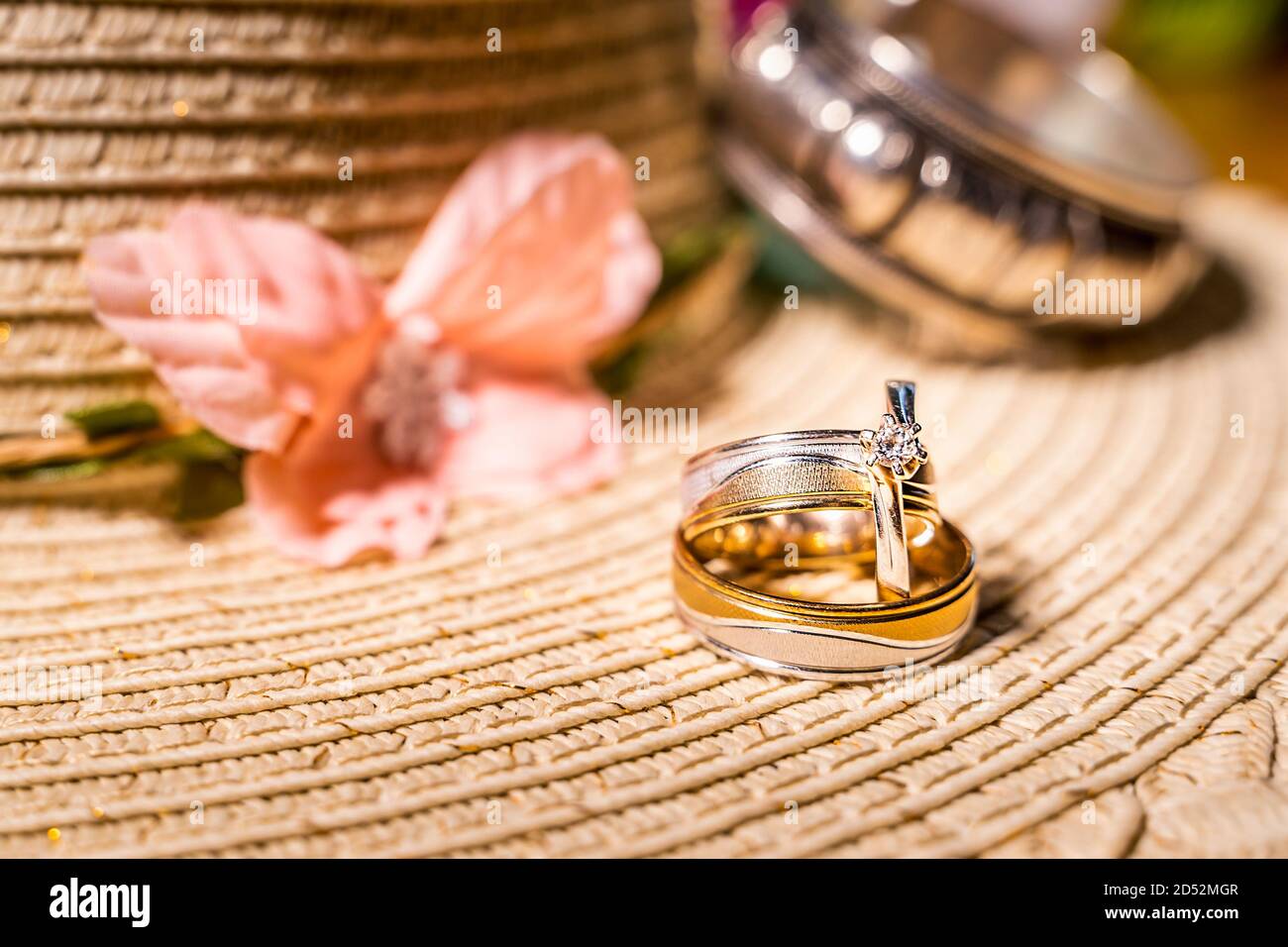 Composizione di due anelli o fasce da sposa e anello di fidanzamento con diamante su sfondo sfocato di cappello di paglia, fiore rosa e bracciale d'argento Foto Stock