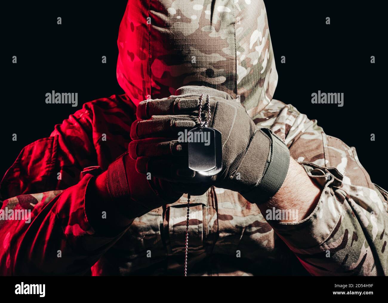 Foto di un soldato militare in uniforme e casco con armatura seduto e che tiene la collana metallica dell'esercito sullo sfondo posteriore con un bagliore rosso. Foto Stock