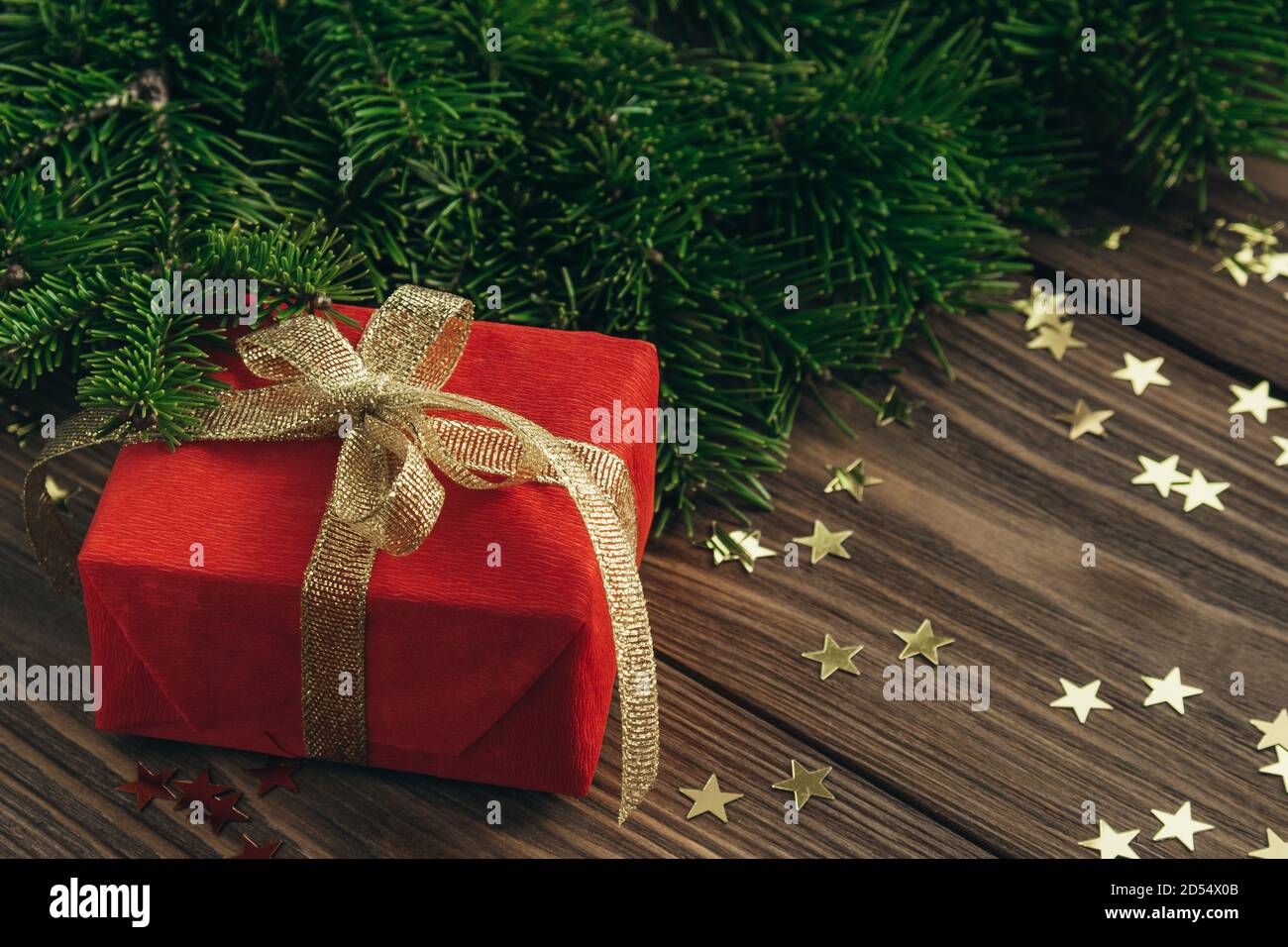 Cartolina natalizia con un regalo rosso con nastro d'oro, rami d'abete e stelle confetti su sfondo ligneo. Messa a fuoco selettiva. Primo piano. Spazio di copia. Foto Stock