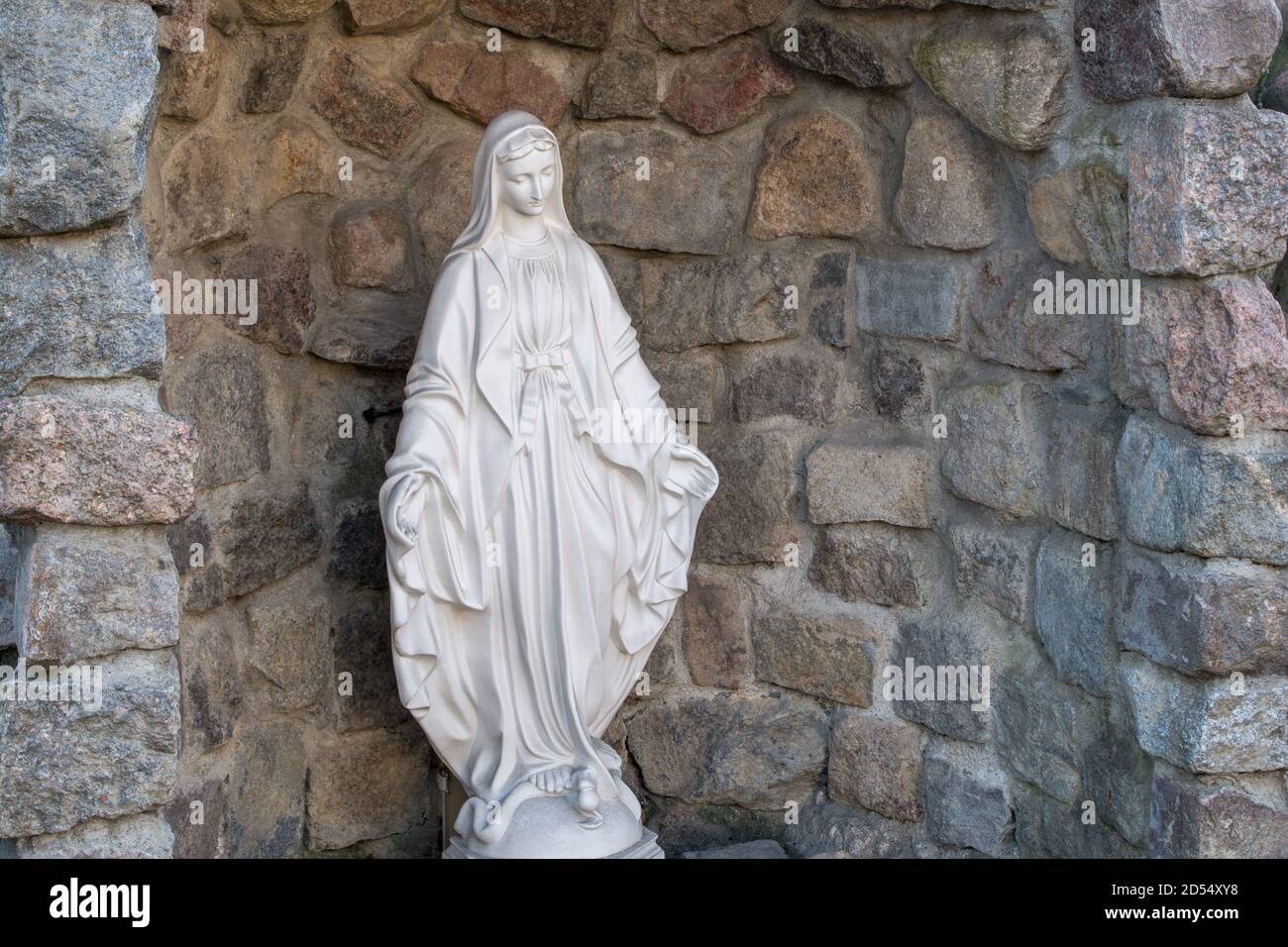 Statua bianca della Vergine Maria in pieghe fluenti di vestiti con testa bombata, braccia distese, piedi calpestanti di serpente di tentatore si trova in grotta Foto Stock
