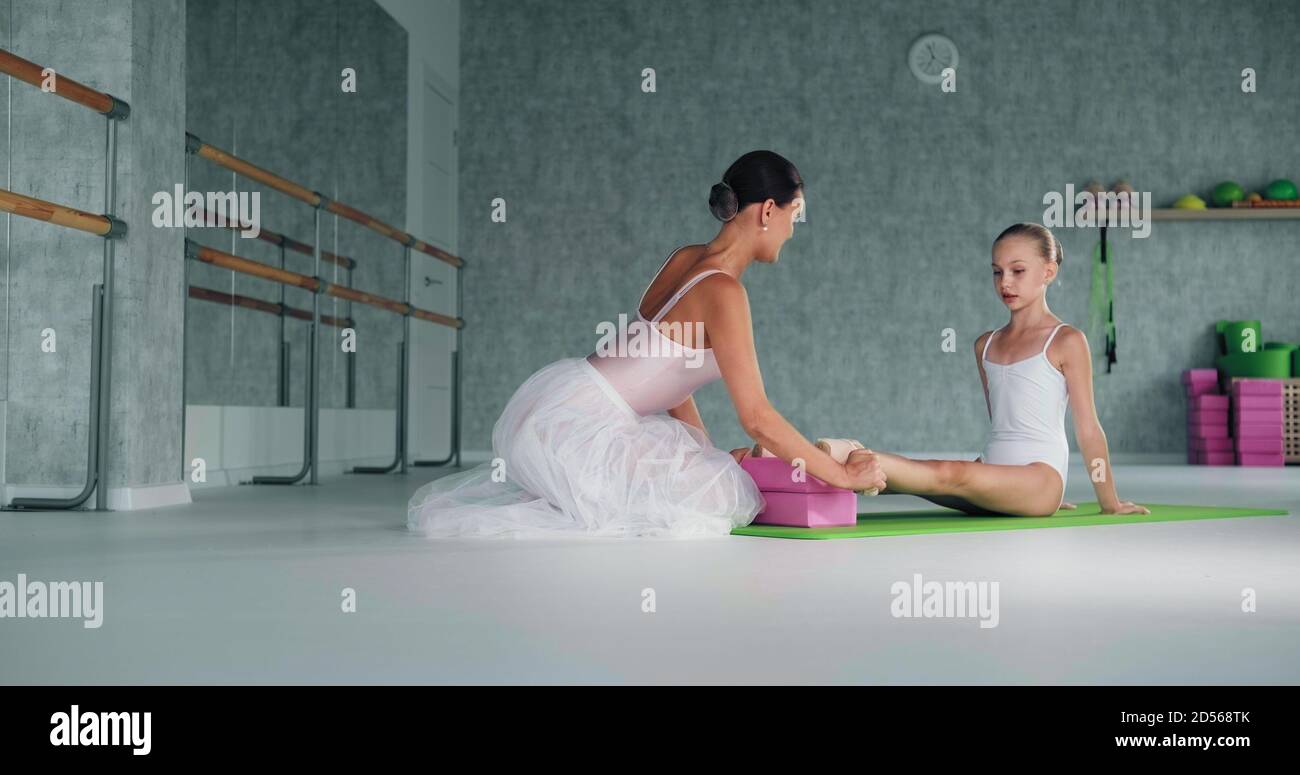 L'addestratore della donna aiuta la ballerina della ragazza della mensa i piedi che si siedono sopra tappetino verde sul pavimento in uno studio luminoso al ballo lezione Foto Stock