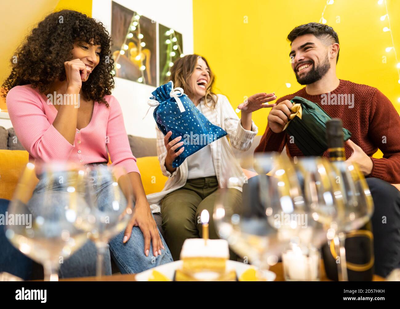 Due giovani e bei amici caucasici danno il loro dono amico nero che celebra il suo compleanno a casa ridendo e scherzando - gara mista a casa par Foto Stock