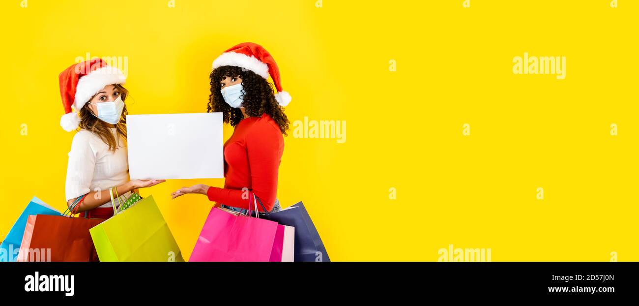 Due giovani donne carine che indossano il cappello di Babbo Natale guardando alla macchina fotografica con gli occhi larghi e la maschera medica, tenendo il segno per lo spazio di copia con molti shopping bag Foto Stock