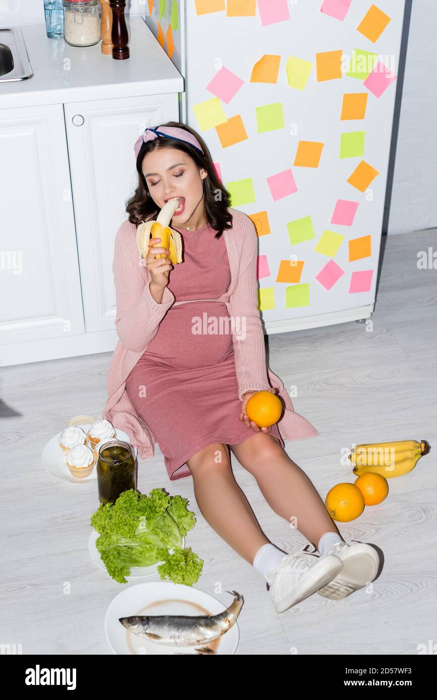 donna incinta che mangia banana vicino al vaso con cetrioli in scatola, cupcakes e arance mentre si siede sul pavimento Foto Stock
