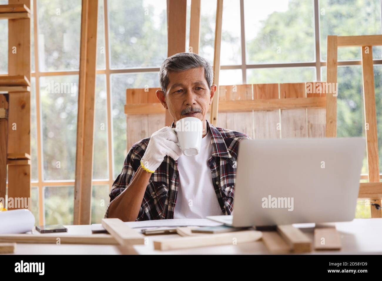 L'uomo anziano beve caffè e lavora con il computer portatile in officina di Falegnameria. Concetto di persone anziane che fanno hobby dopo il pensionamento. Foto Stock