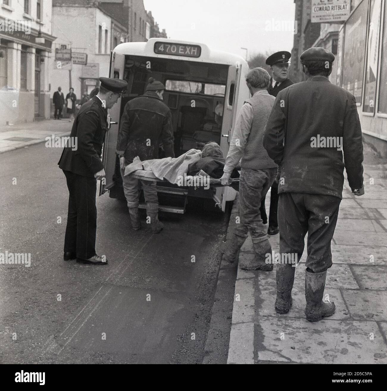 Anni '60, storico, fuori in una strada, un lavoratore ferito su una barella che viene messo nella parte posteriore di un'ambulanza dai suoi colleghi di lavoro nei loro vestiti fangosi e stivali, con personale maschile in uniforme del London Ambulance Service assistere con le porte, Londra sud-est, Inghilterra, Regno Unito. In quest'epoca, gli inifessi "autisti di ambulanza" hanno preso i pazienti e li hanno direttamente ricoverati in ospedale, a differenza dei tempi moderni in cui il personale dell'ambulanza, molti con qualifiche scientifiche paramediche, spesso tratta i pazienti in scena di incidenti. Foto Stock