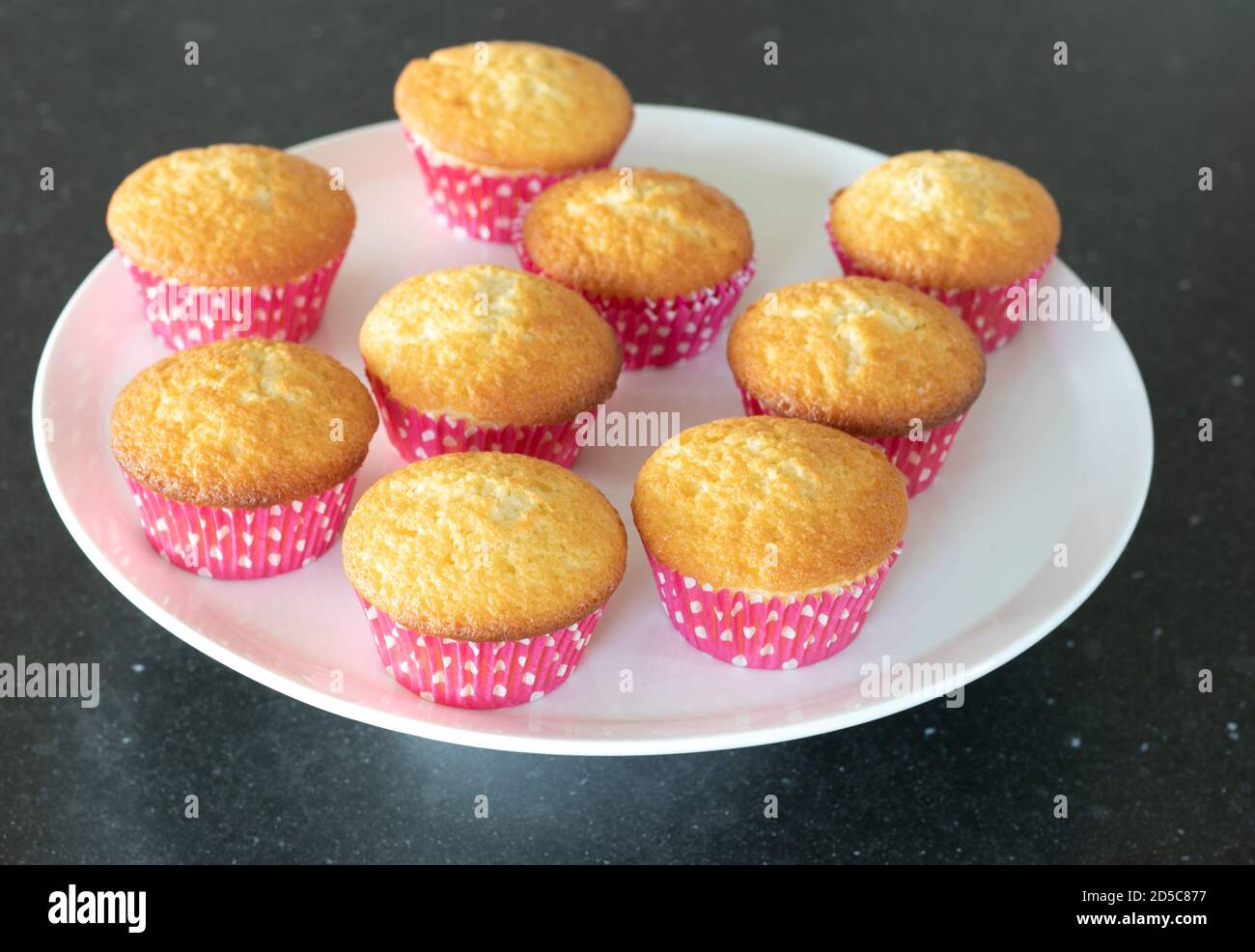 Plein cupcakes cotto in casa in un piatto bianco, vista dall'alto su uno sfondo grigio scuro, tazze rosa con puntini bianchi Foto Stock