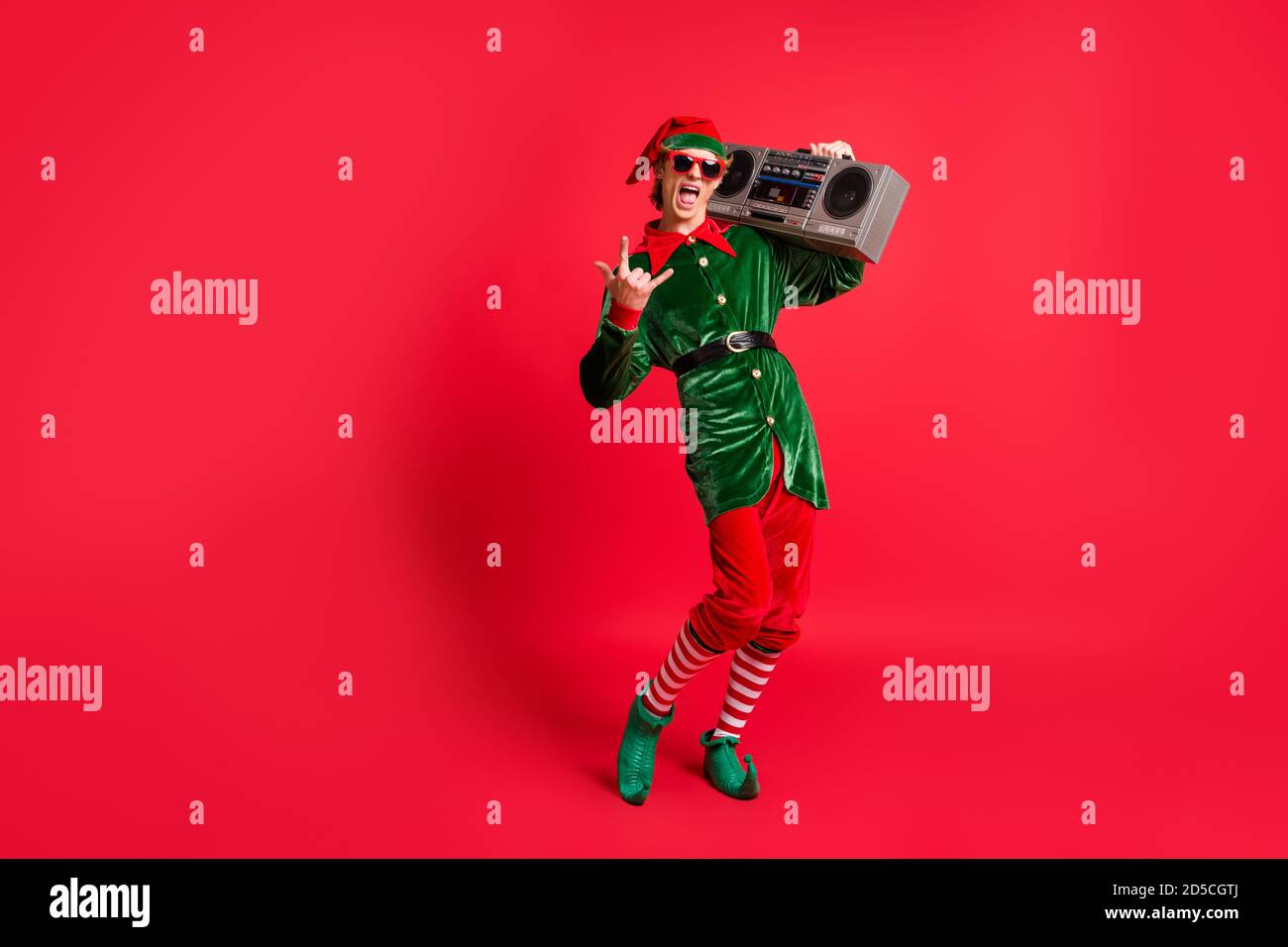 Foto a dimensione intera di pazzo elfo tenere braccio box ascoltare le canzoni x-mas mostrano il simbolo a corna indossa un costume verde isolato sfondo di colore rosso Foto Stock