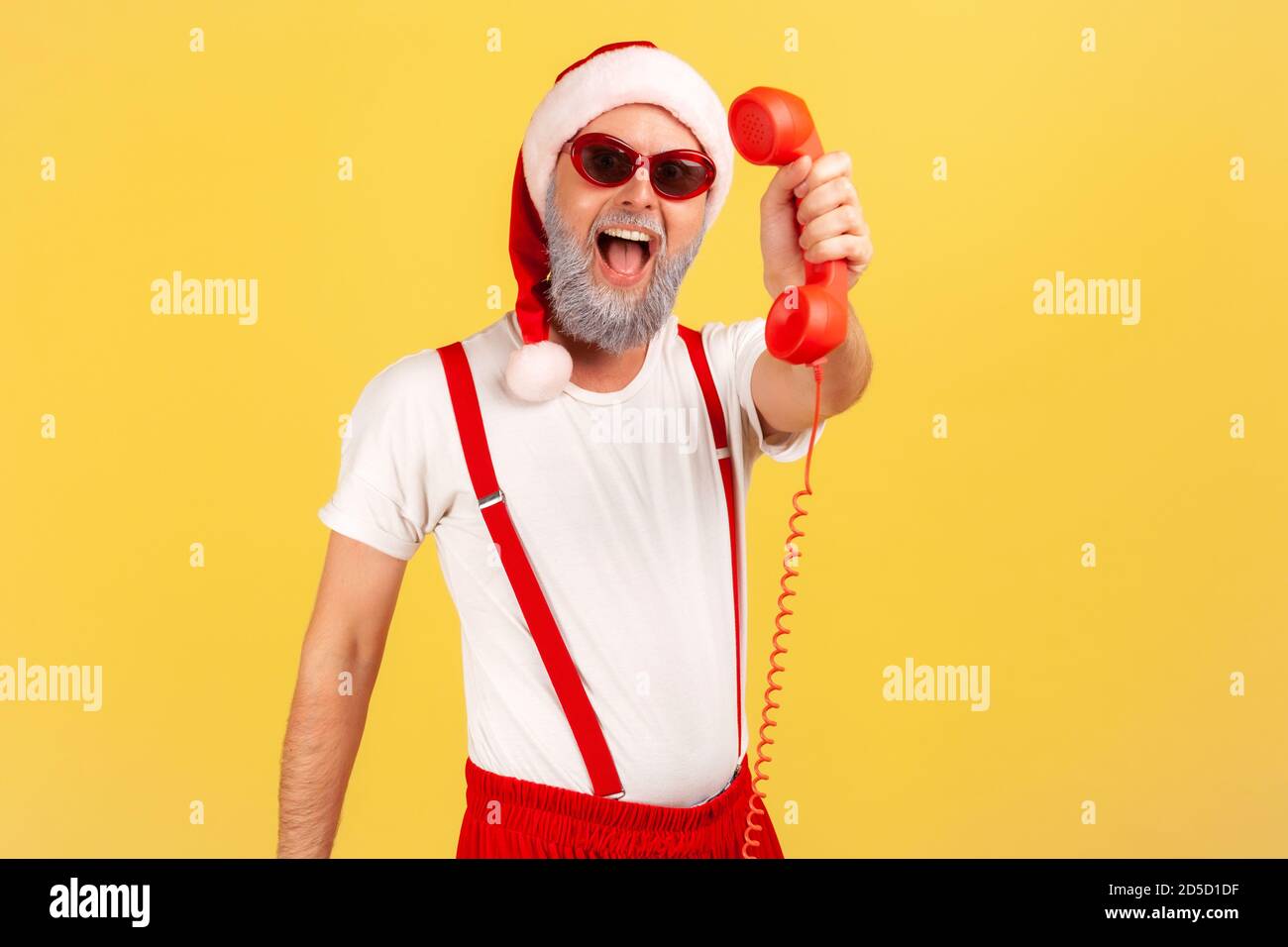 Eccitato felice uomo con bearded grigio in occhiali da sole e cappello di babbo natale che tiene e mostra telefono fisso, chiamando per congratularsi con le casse. IO Foto Stock