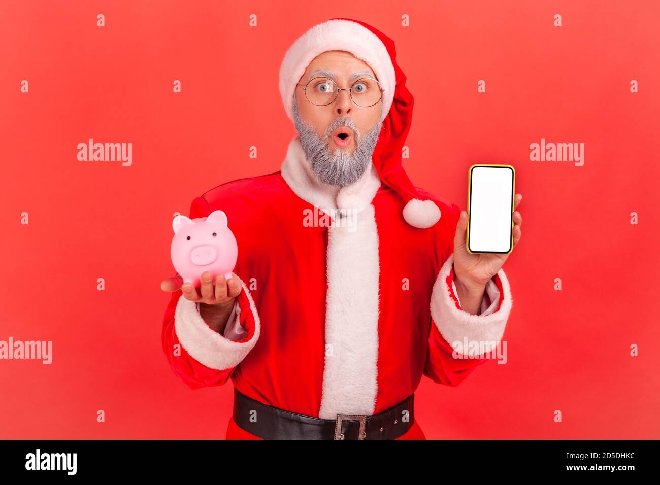 Scioccato sorpreso babbo natale tenere smartphone con schermo vuoto e piggy banca, cashback, portafoglio online. Studio al coperto isolato su sfondo rosso Foto Stock