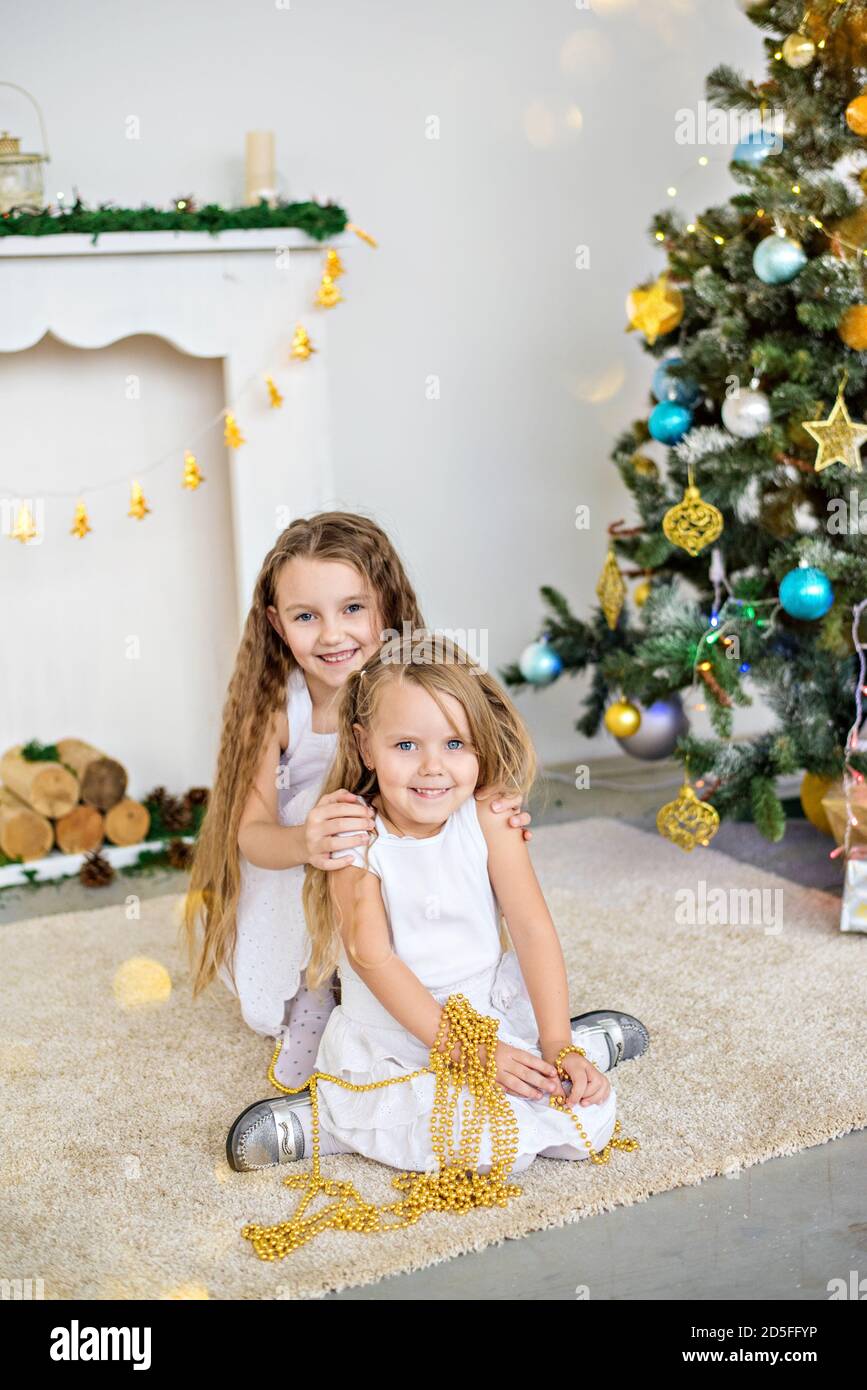 Due ragazze bionde piccole in abiti bianchi stanno giocando vicino al camino e l'albero di Natale con le scatole. Le sorelle hanno divertimento, abbraccio, ridono, scoprono i regali Foto Stock