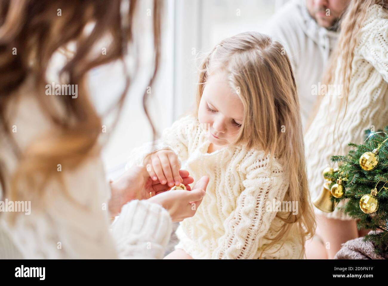 Una madre bionda e una figlia in bianco maglia pullover decorare un piccolo albero di Natale con giocattoli d'oro. Una giovane donna mostra nelle mani la ragazza anno nuovo Foto Stock
