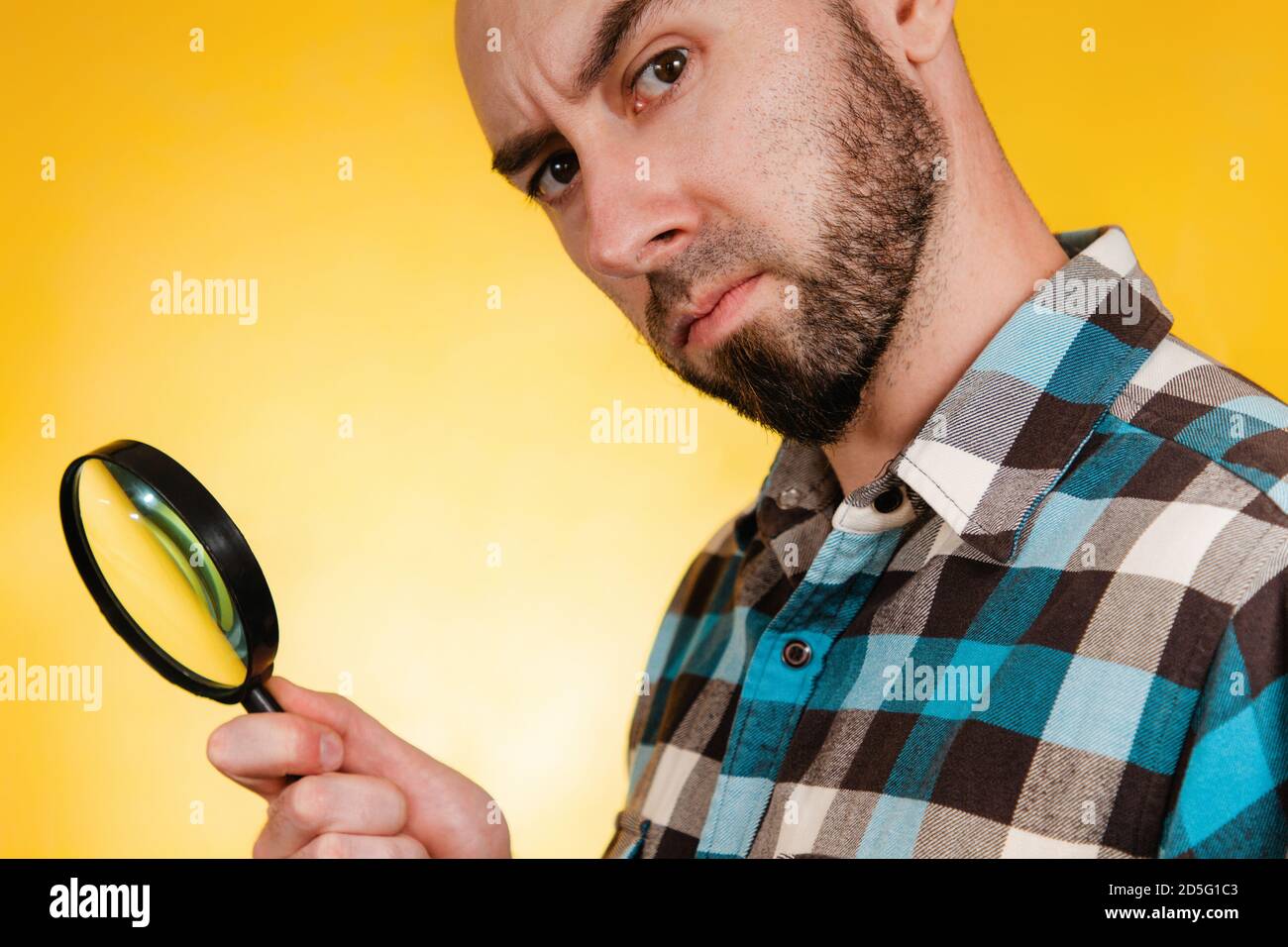 Il concetto di trovare e risolvere i problemi. Un uomo calvo con la barba, vestito con una camicia blu a forma di plaid, che tiene una lente d'ingrandimento e guarda attentamente. Foto Stock