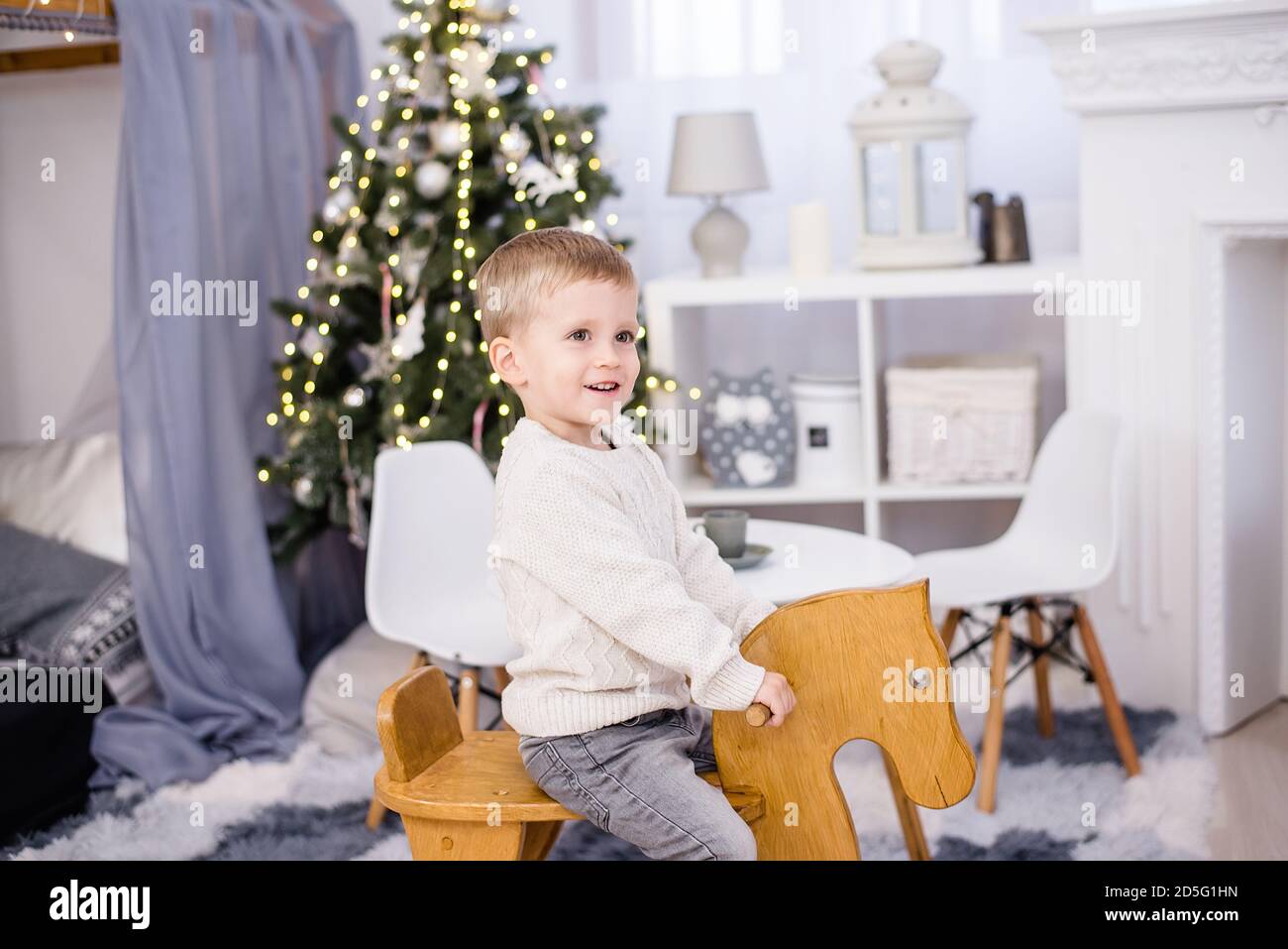Felice bambino biondo in un pullover bianco lavorato a maglia e. i jeans  cavalcano un cavallo in legno a dondolo nella stanza dei bambini Accanto ad  un albero di Natale con ghirlande