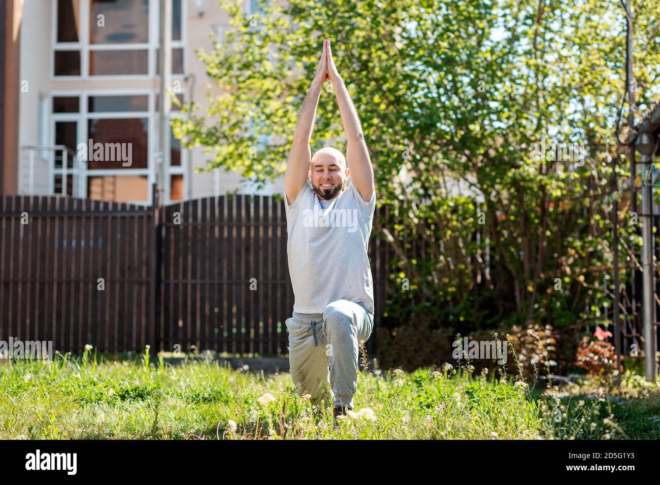Un giovane uomo felice in abiti sportivi sta facendo yoga sull'erba nel suo cortile. All'aperto. Il concetto di autoisolamento e sport. Foto Stock