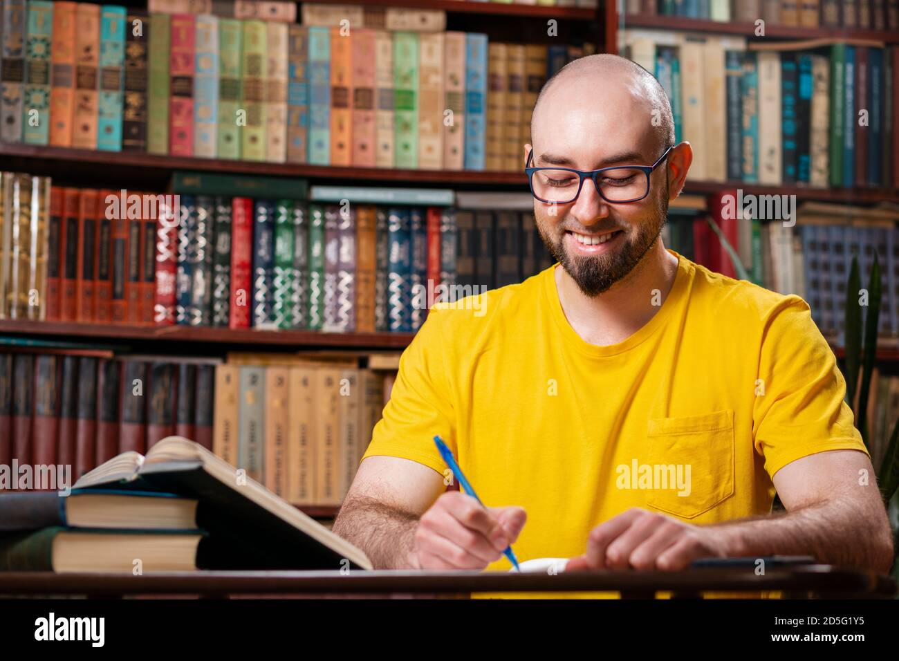 Un uomo calvo felice, bearded con gli occhiali sta scrivendo qualcosa in un taccuino. Ci sono librerie sullo sfondo. Concetto di apprendimento a distanza ed e Foto Stock