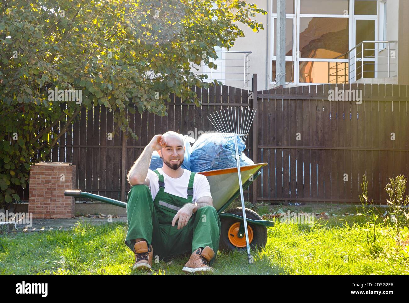 Il giardiniere nell'uniforme verde sta poggiando sull'erba. Sorriso e buon umore. Il sole splende sul lato destro. Foto Stock