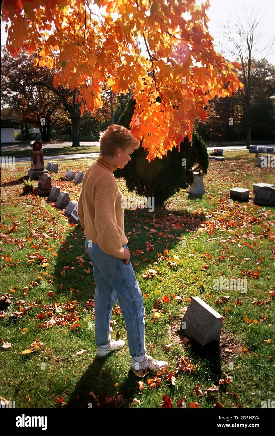 La donna visita un luogo della tomba di una persona amata in un cimitero con albero colorato sullo sfondo Foto Stock