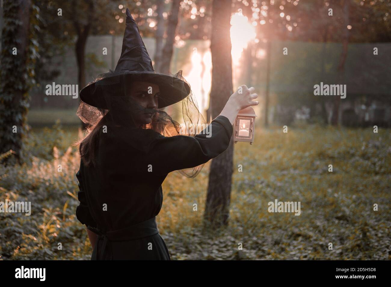 Giovane donna in abito scuro e cappello di strega tiene lanterna con candele in mano e illumina la foresta. Costume da festa di Halloween. Parco con autum Foto Stock