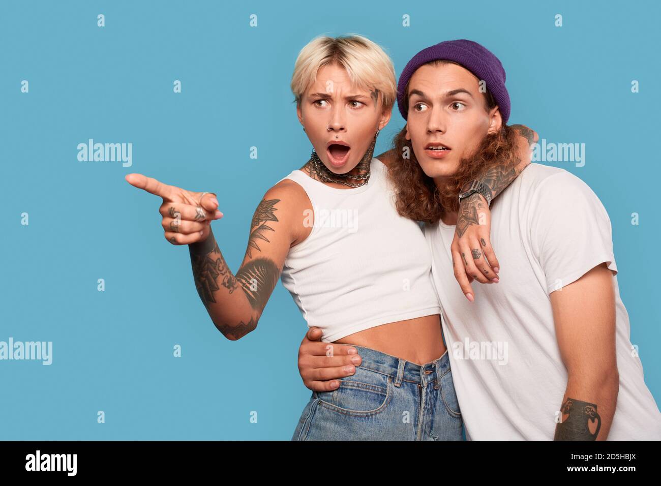 Stile libero. Giovane coppia tatuata in piedi isolata su fidanzata blu hugging boyfriend che punta da parte scioccato Foto Stock