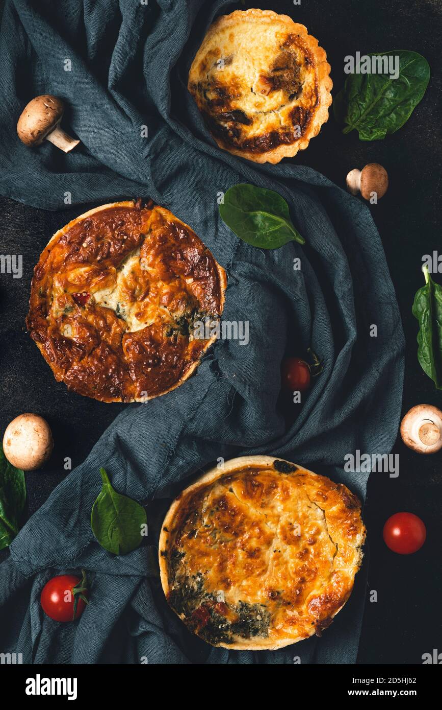 Still life con torte fatte in casa quiche lorraine, tessuti scuri, spinaci, pomodori e funghi su tavola nera. Vista dall'alto. Piatto Foto Stock