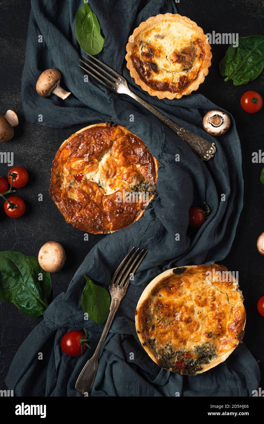 Pollo, funghi, spinaci e pomodori torta quiche con posate su tessuto scuro su tavola nera. Vita morta. Disposizione piatta. Vista dall'alto Foto Stock