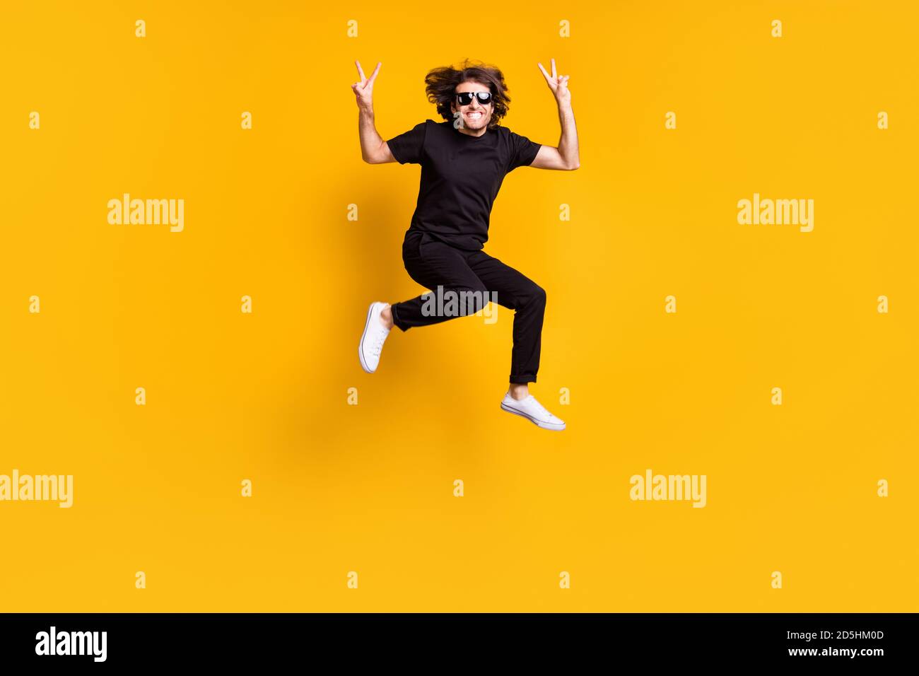Ritratto di uomo che salta in mostra a tutta lunghezza gesto v-sign con entrambe le mani in outfit nero sunglass isolato di colore giallo brillante Foto Stock