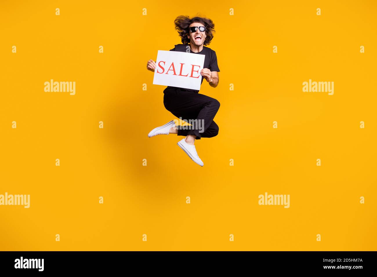 Ritratto di un uomo che salta in alto gridando a tutta lunghezza mantenere la vendita della tabella che indossa i vestiti neri sunglass isolato su luminoso colore giallo Foto Stock