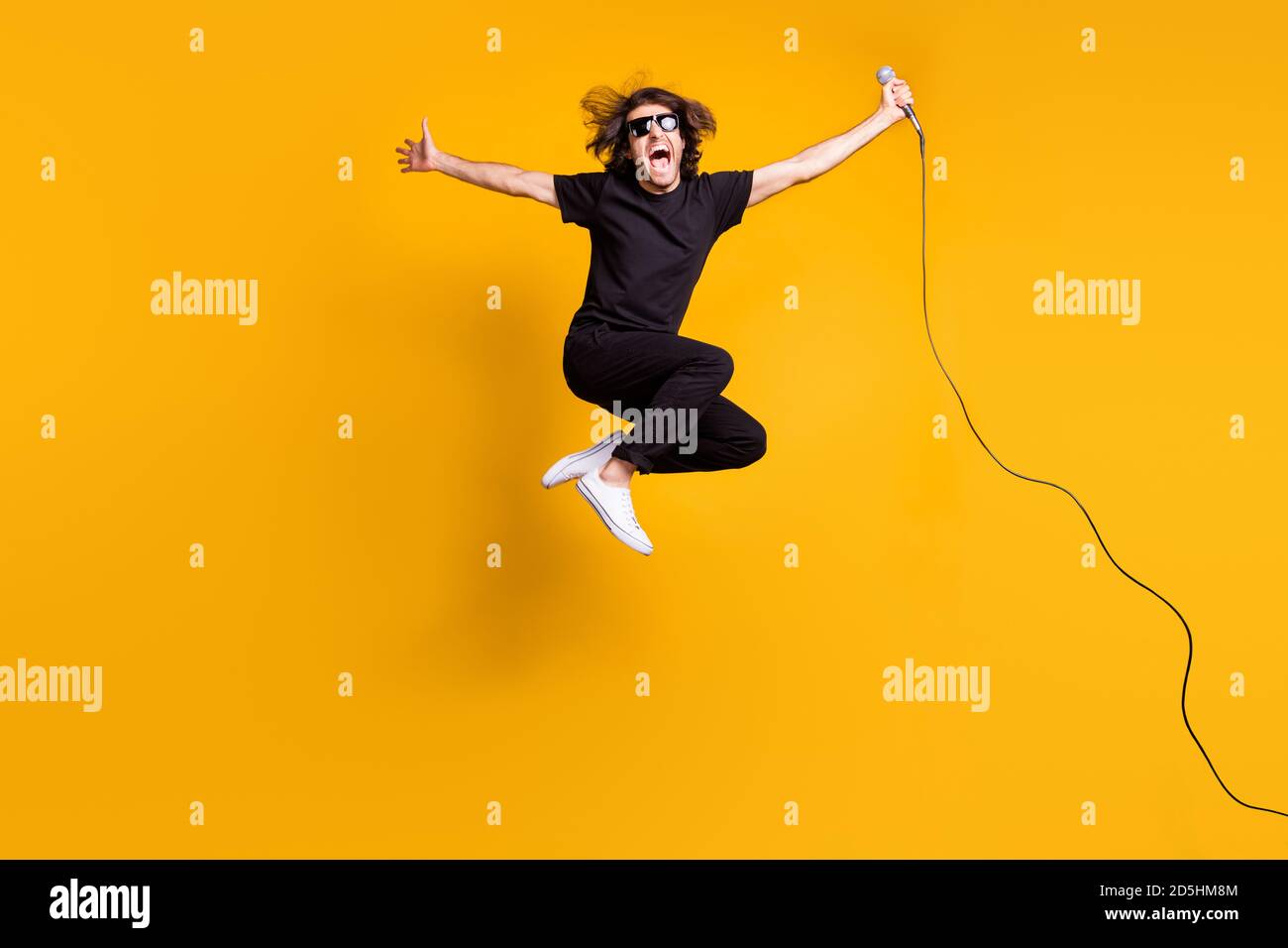 Ritratto di uomo che indossa abiti neri a tutta lunghezza la sunglass salta ad alto grido mantenendo il microfono isolato sul giallo brillante colore Foto Stock