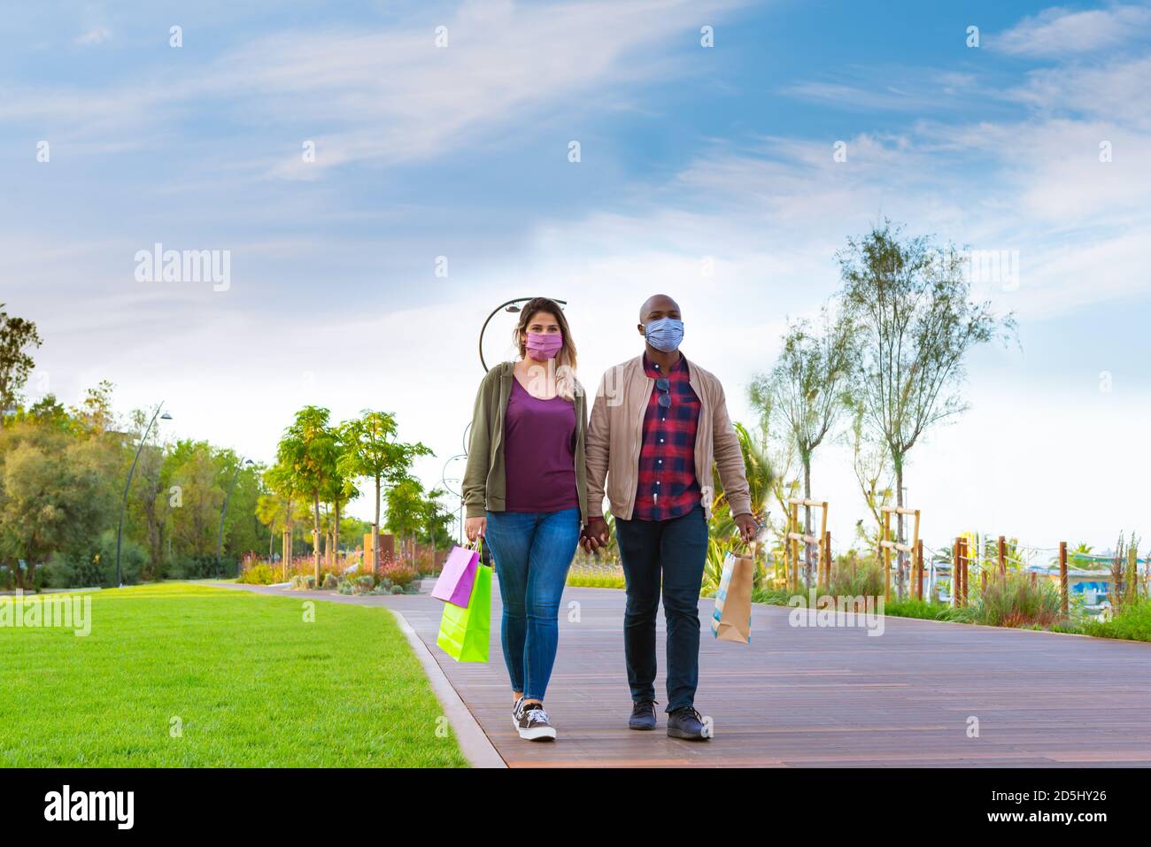 attraente coppia interracial che indossa maschera facciale che tiene le mani con lo shopping borse di carta mentre camminando in un parco in autumn.concept circa il nuovo normale Foto Stock