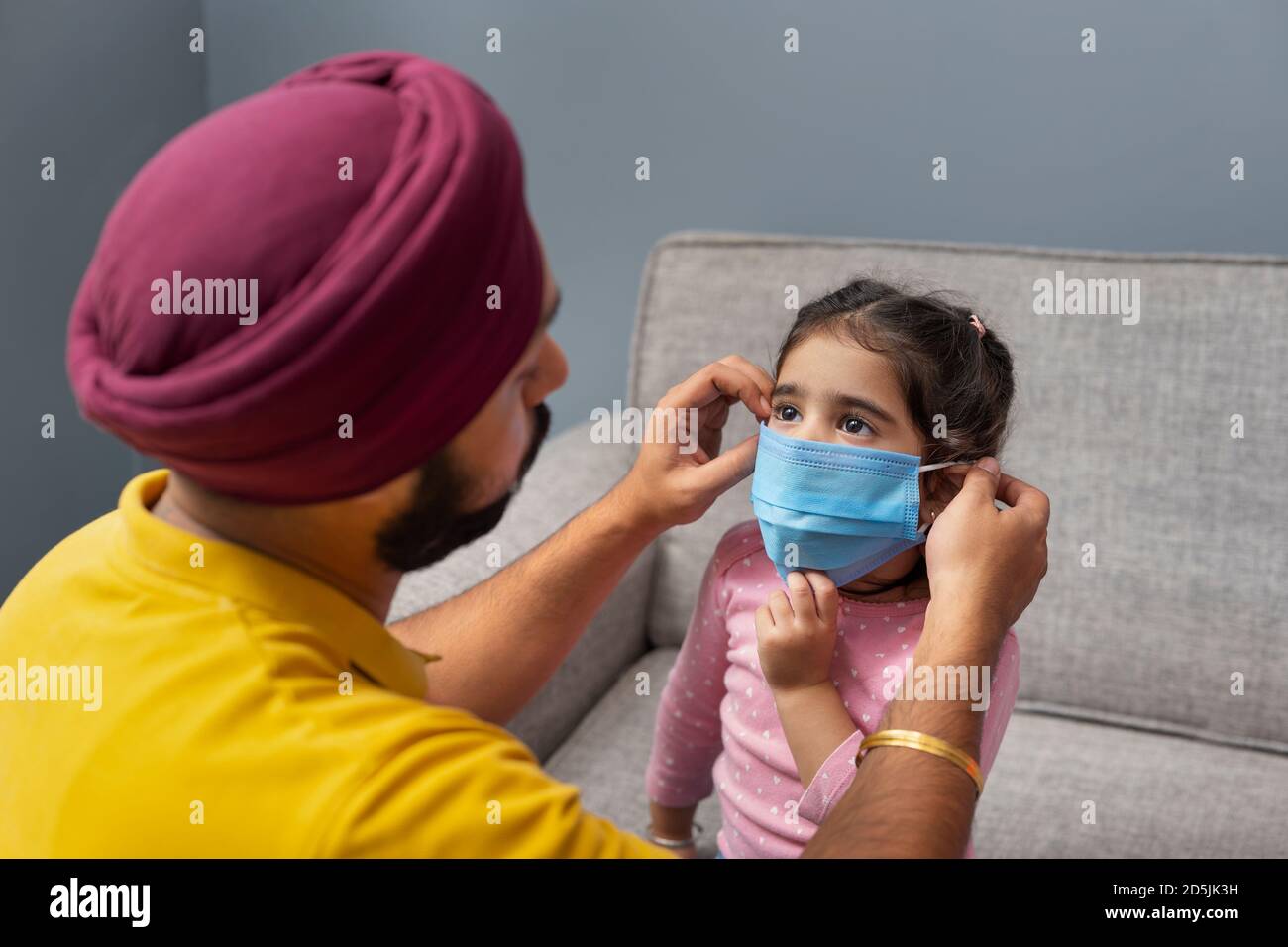 UNA PICCOLA RAGAZZA SIKH CHE VIENE AIUTATA DAL PADRE A INDOSSARE MASCHERA PRIMA DI USCIRE DALLA CASA Foto Stock