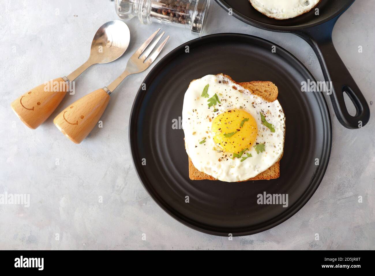 Uovo fritto su pane integrale con sale e pepe per la colazione classica. Uovo soleggiato con pane marrone su piatto nero su tavolo di legno. Foto Stock