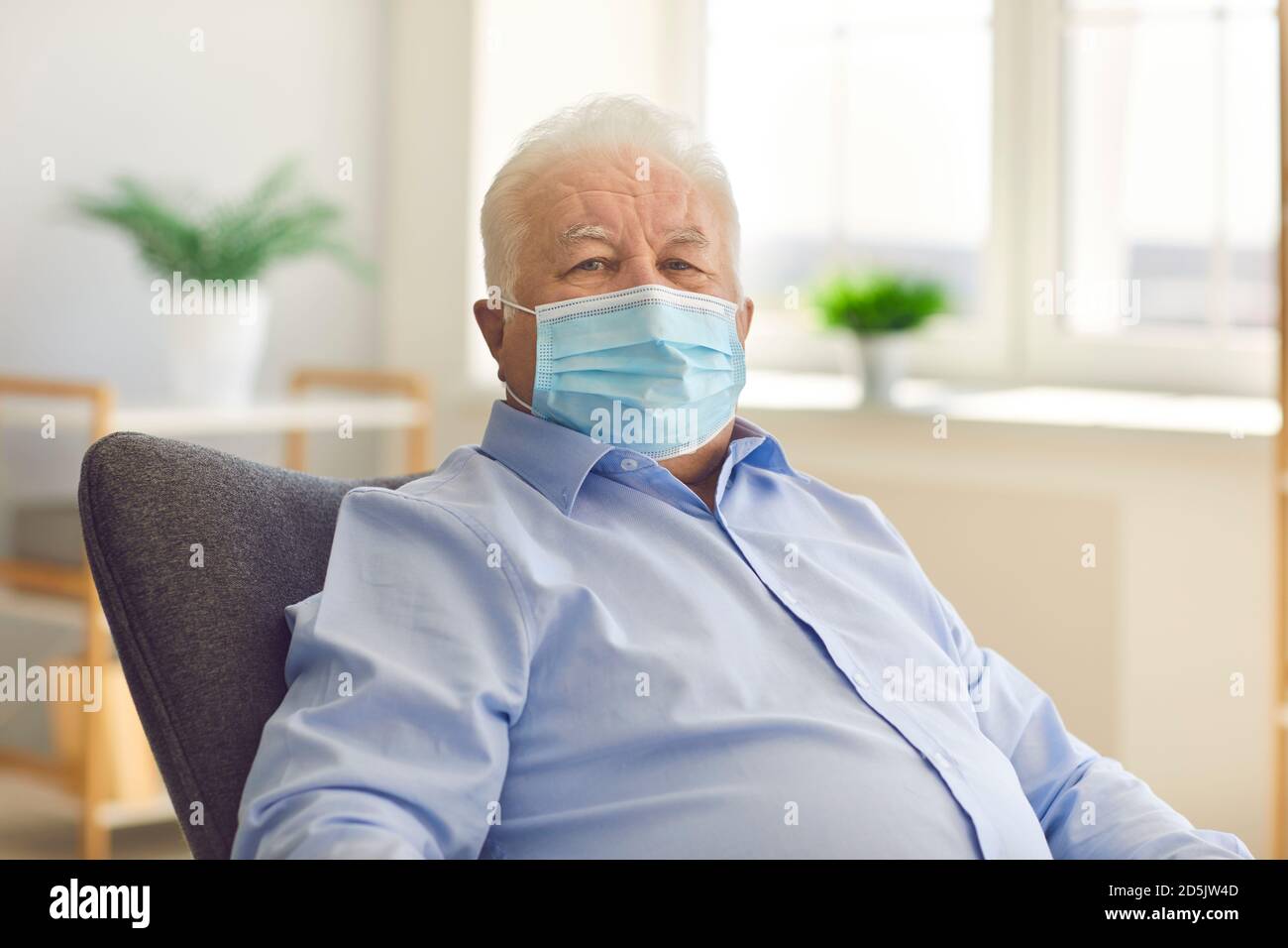 Uomo anziano serio che indossa una maschera medica seduta in poltrona guardando la fotocamera Foto Stock