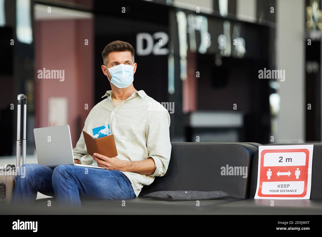 Giovane uomo in maschera protettiva con bagaglio e biglietti seduti in aeroporto durante la pandemia Foto Stock
