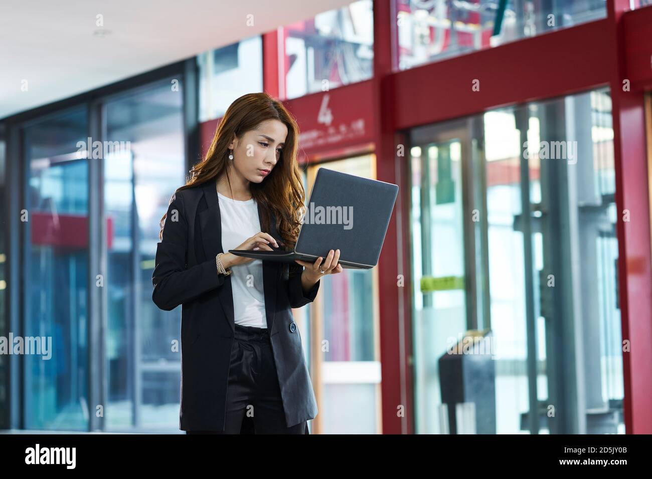 donna d'affari asiatica che controlla la posta elettronica utilizzando un computer portatile mentre si cammina lobby aziendale Foto Stock