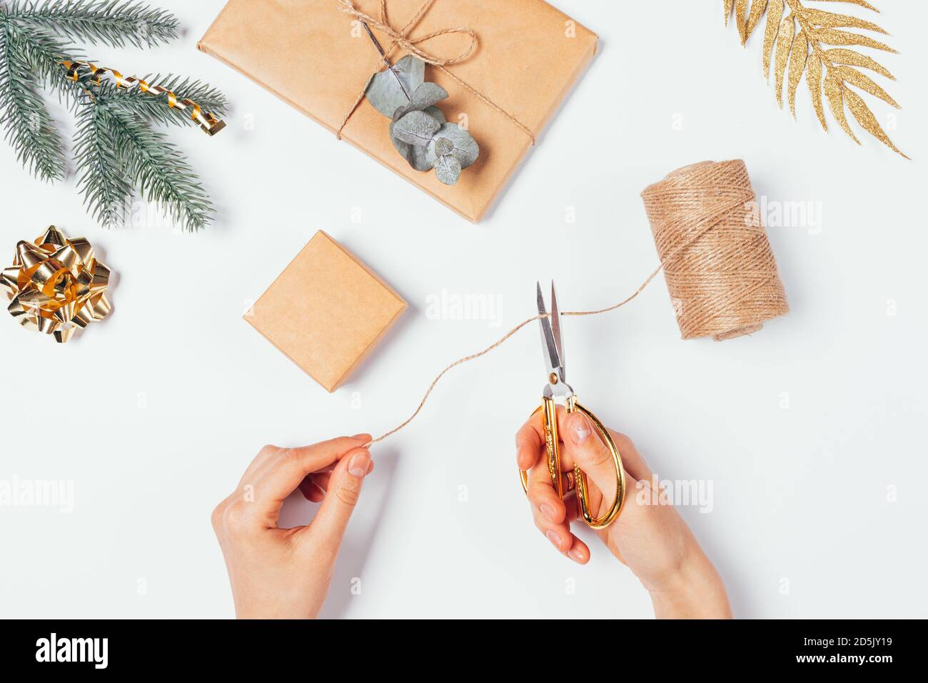 Le mani femminili tengono le forbici tagliando lo spago per fare l'imballaggio fatto in casa per i regali di Natale vicino alle scatole del mestiere marrone, le decorazioni dorate, l'abete e l'euca Foto Stock