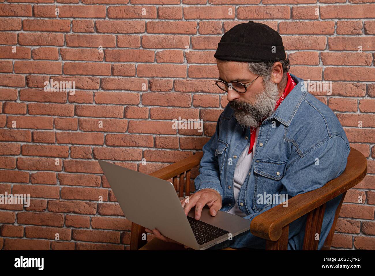 Vista laterale ritratto di uomo bearded in camicia di denim seduta su sedia di legno che lavora su laptop. Foto Stock