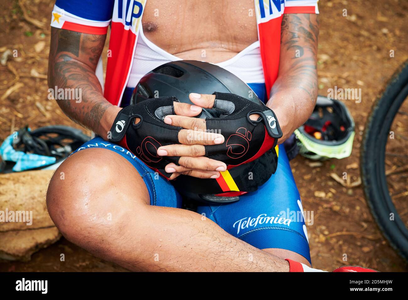 Vista ravvicinata di un ciclista tatuato in abbigliamento da corsa sta riposando e tenendo il casco con le mani, indossando guanti da bicicletta Foto Stock