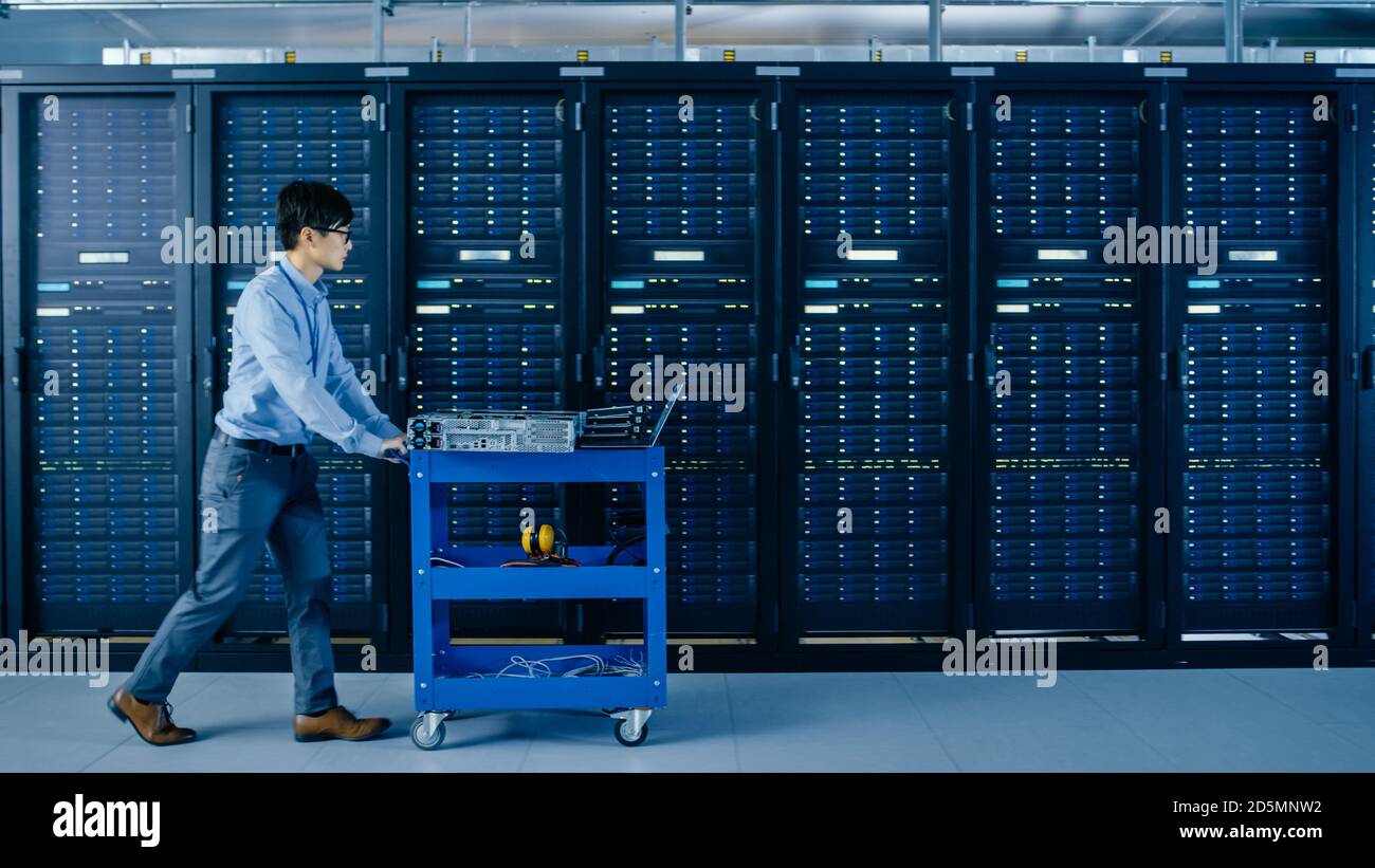 Nel moderno data center: Il tecnico IT spinge il carrello accanto a rack server e cabinet per installare nuovo hardware per l'aggiornamento pianificato del sistema e le apparecchiature Foto Stock