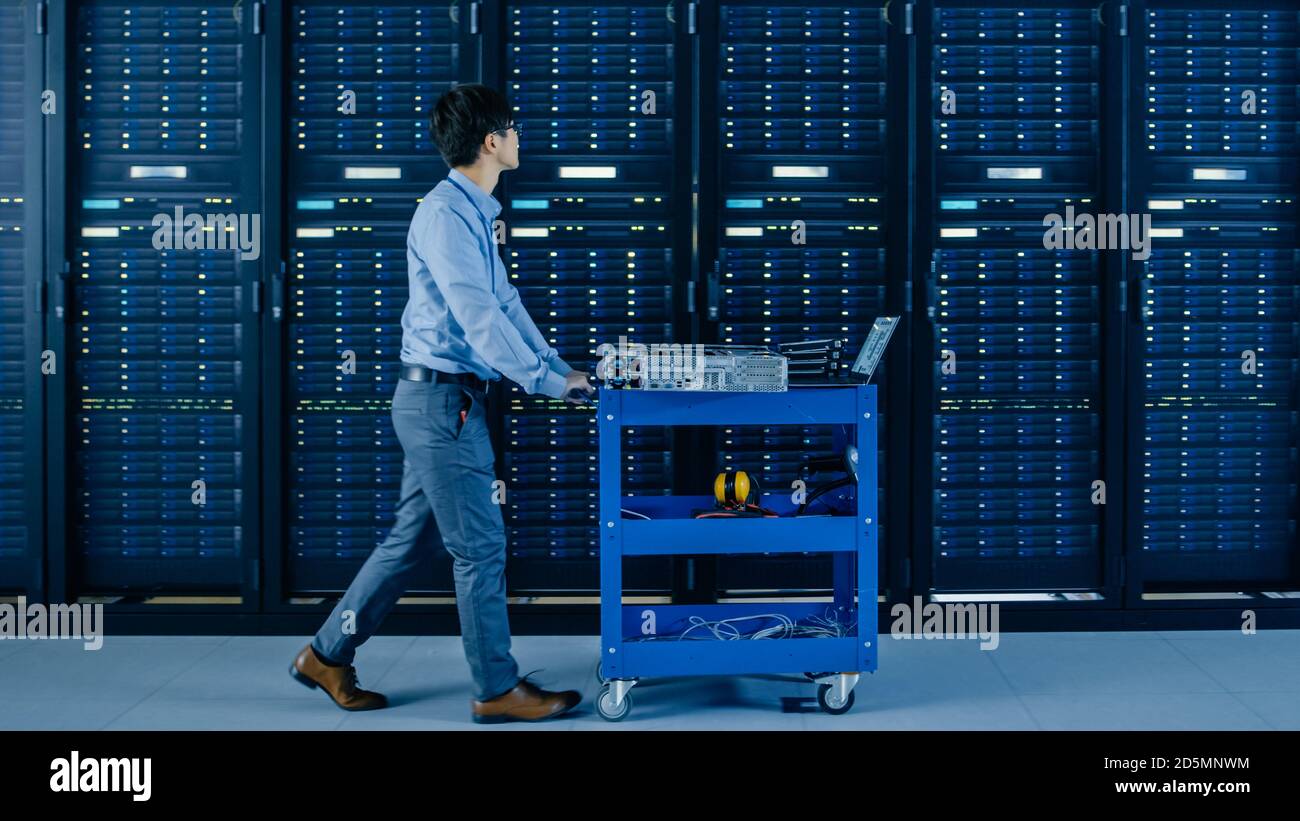 Nel moderno data center: Il tecnico IT spinge il carrello accanto a rack server e cabinet per installare nuovo hardware per l'aggiornamento pianificato del sistema e le apparecchiature Foto Stock