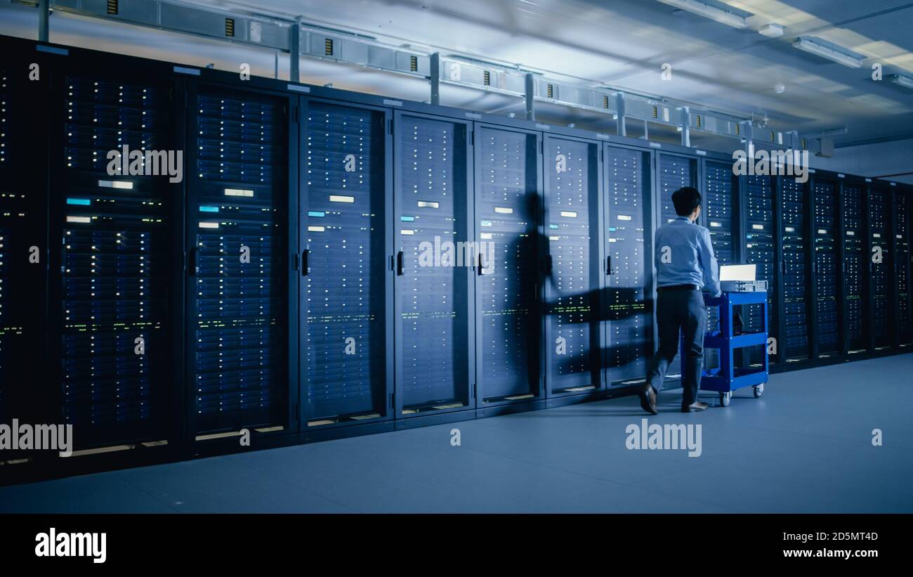 Nel moderno data center: Tecnico IT che esegue la manutenzione e la diagnostica. Spinge il carrello accanto ai rack server. Aggiornamento pianificato delle apparecchiature di sistema e. Foto Stock